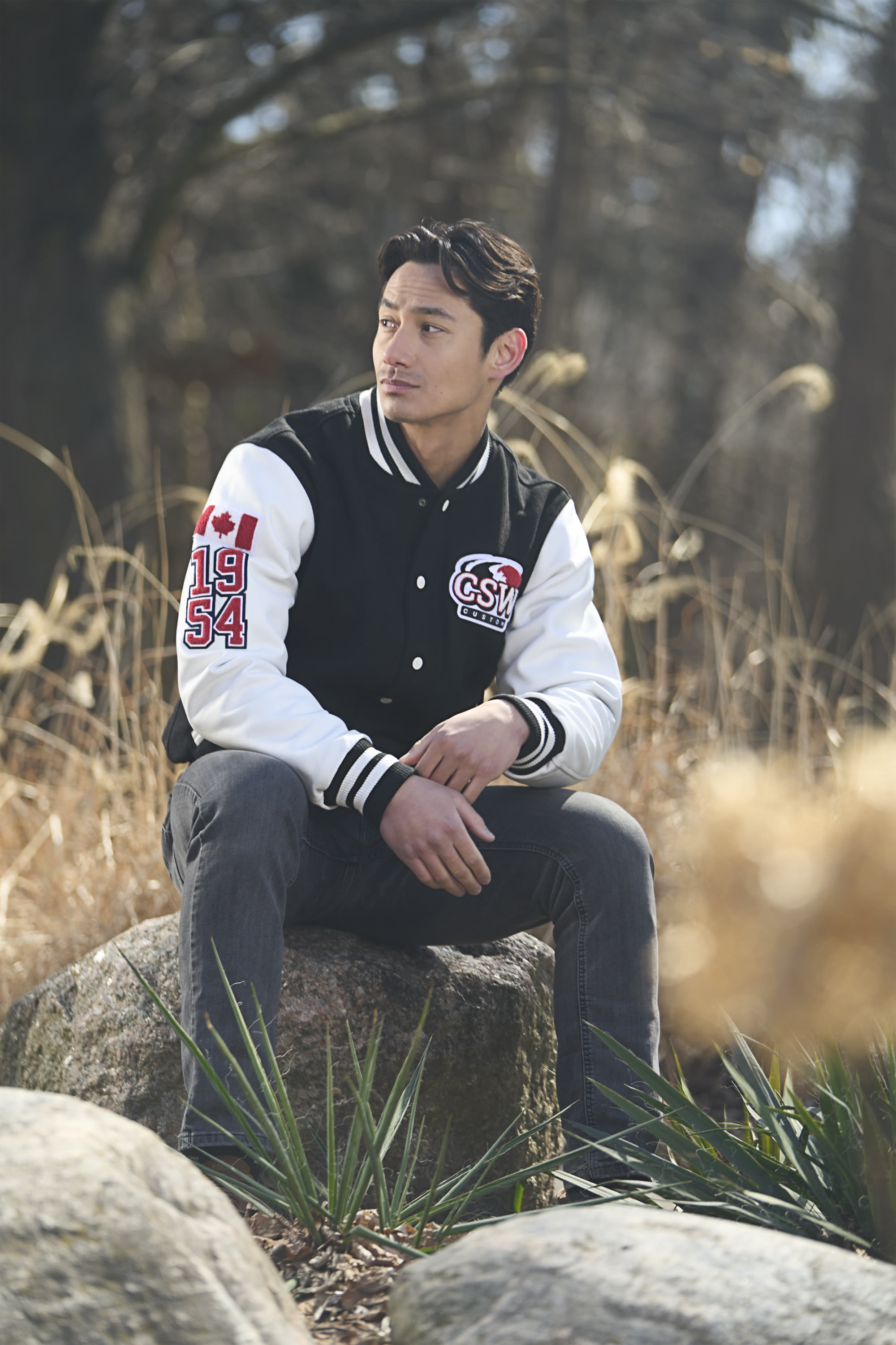 Model in a black and white Canada Sportswear varsity jacket seated on a rock in dry winter grass - catalog photography by Fisheye Studio Toronto